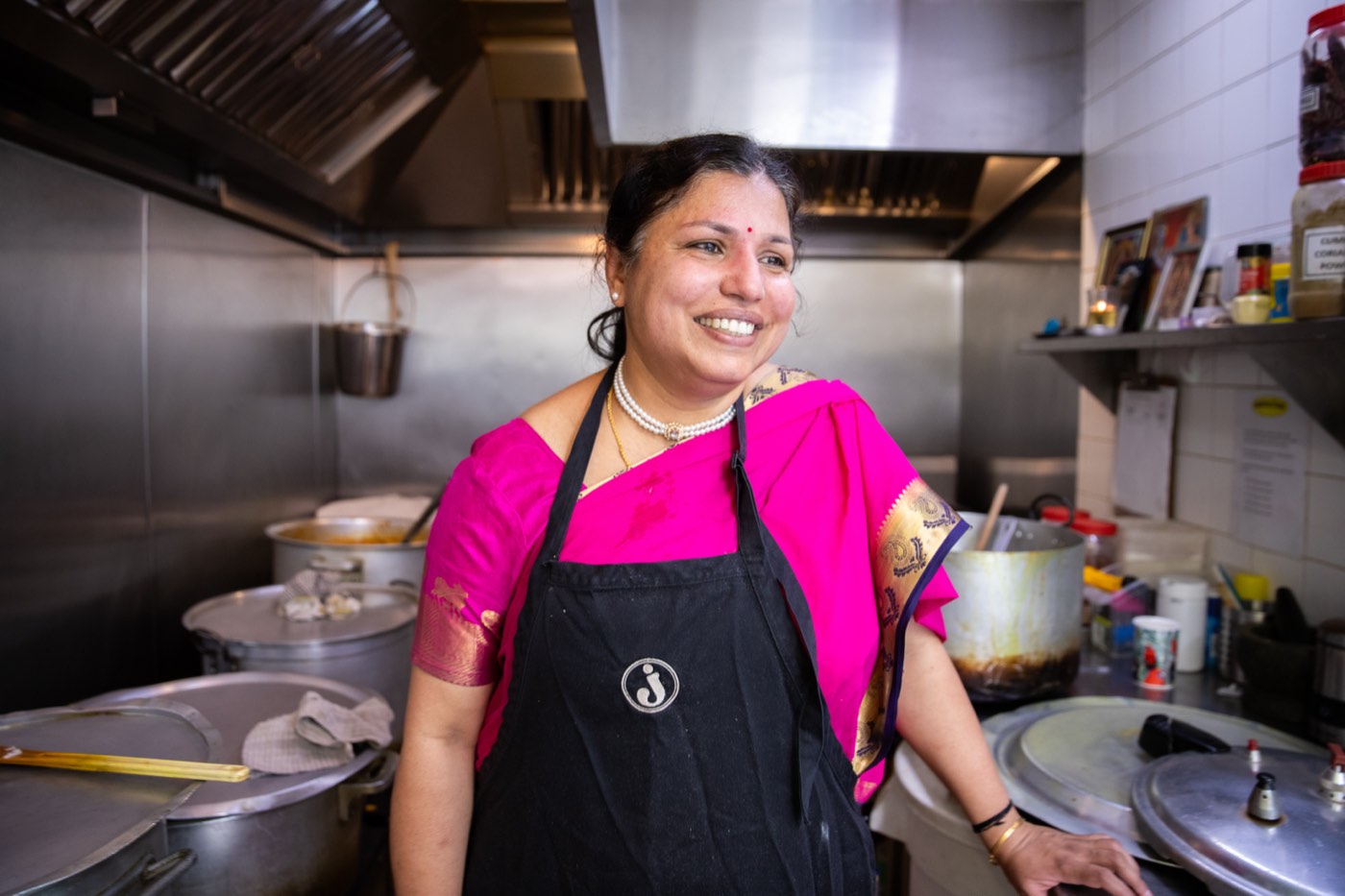 A volunteer in a pink sari and a black apron, smiling in the ashram kitchen beside the large cooking pots.